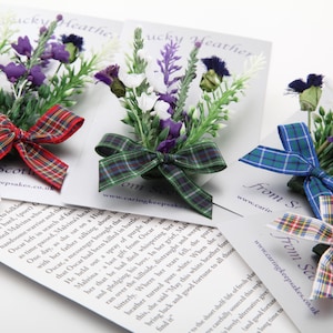 May include: Three Scottish heather flower brooches with tartan ribbon bows. The brooches are on white cards with the text "From Scotland" and a website address.
