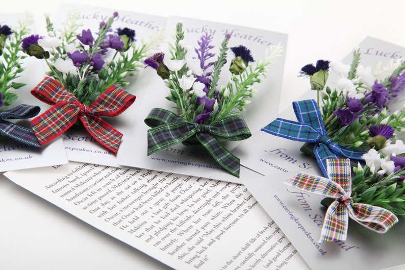 May include: Three Scottish heather flower brooches with tartan ribbon bows. The brooches are on white cards with the text "From Scotland" and a website address.