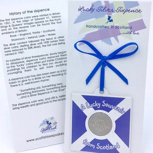 May include: A silver sixpence coin from Scotland, displayed on a blue and white Scottish flag design with the text "A Lucky Souvenir from Scotland". The coin is surrounded by a blue ribbon tied in a bow. The back of the card has a history of the sixpence coin and the text "Lucky Silver Sixpence Handcrafted in Scotland...with love".
