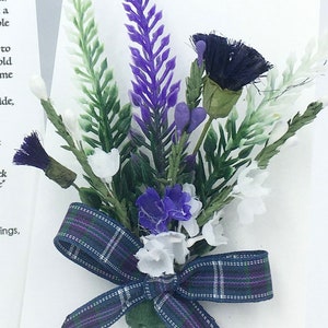 May include: A small bouquet of purple and white flowers with a blue and green plaid ribbon. The flowers are arranged in a cluster and tied together with the ribbon. The bouquet is on a white background.