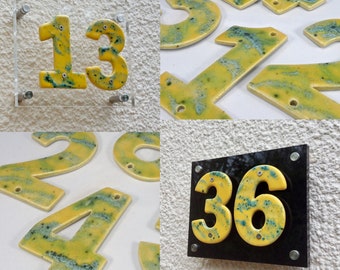 Może przedstawiać: Zestaw żółtych ceramicznych numerów domowych z zielonymi i niebieskimi plamkami. Numery to 1, 2, 3, 4, 6 i 13. Numery są zamontowane na białej ścianie.