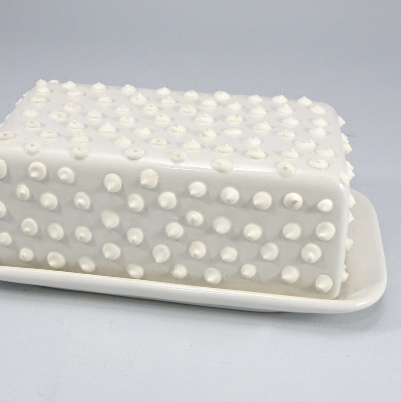 Puede incluir: Una mantequera de cer&aacute;mica blanca con tapa y base rectangulares. La tapa est&aacute; cubierta de peque&ntilde;as protuberancias redondeadas en relieve. El plato est&aacute; dise&ntilde;ado para contener un tama&ntilde;o de mantequilla est&aacute;ndar. El dise&ntilde;o general es minimalista y moderno.