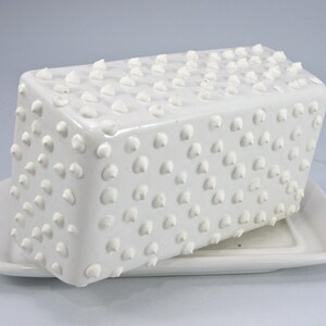 Puede incluir: Una mantequera de cer&aacute;mica blanca con tapa y base rectangulares. La tapa est&aacute; cubierta de peque&ntilde;as protuberancias redondeadas en relieve. La base tiene una forma rectangular simple. El plato est&aacute; dise&ntilde;ado para contener mantequilla.