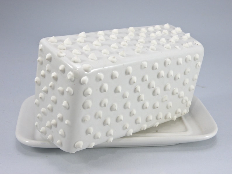 Puede incluir: Una mantequera de cer&aacute;mica blanca con tapa y base rectangulares. La tapa est&aacute; cubierta de peque&ntilde;as protuberancias redondeadas en relieve. La base tiene una forma rectangular simple. El plato est&aacute; dise&ntilde;ado para contener mantequilla.