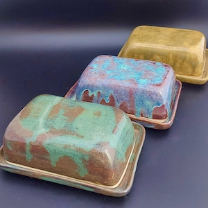 May include: Three ceramic butter dishes in different colours and patterns. The dishes are glazed in shades of brown, green, blue, and yellow. The dishes are all rectangular and have a raised rim.