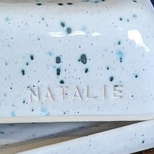 May include: White ceramic butter dish with blue and black speckles. The dish has a raised rim and the name "NATALIE" is embossed on the front.