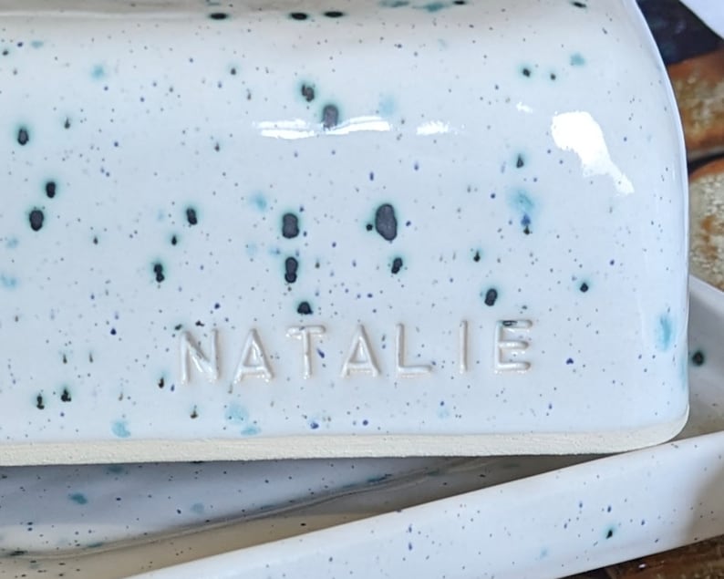 May include: White ceramic butter dish with blue and black speckles. The dish has a raised rim and the name "NATALIE" is embossed on the front.