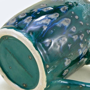May include: A green ceramic mug with a speckled glaze and a blue and silver swirl design. The bottom of the mug has the website "www.Domspottery.com" printed on it.