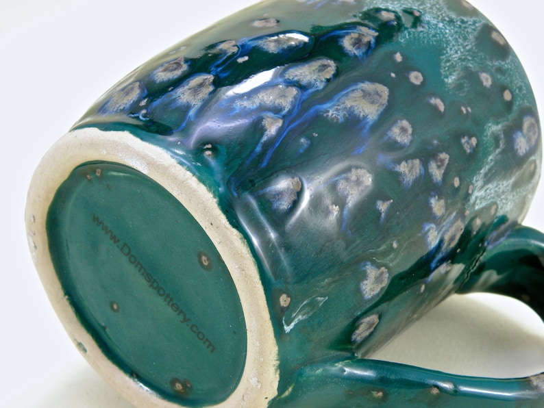 May include: A green ceramic mug with a speckled glaze and a blue and silver swirl design. The bottom of the mug has the website "www.Domspottery.com" printed on it.