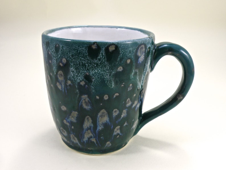May include: A green ceramic mug with a white interior and a speckled glaze. The mug has a rounded body and a handle.