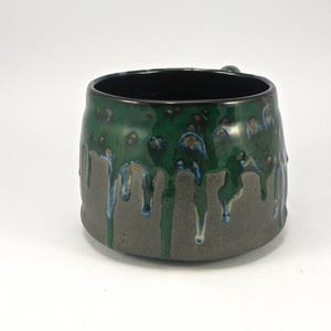 May include: A green and gray ceramic mug with a drip glaze design. The mug has a wide, rounded shape and a slightly flared lip.