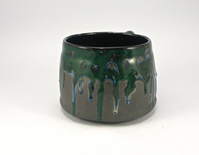 May include: A green and gray ceramic mug with a drip glaze design. The mug has a wide, rounded shape and a slightly flared lip.
