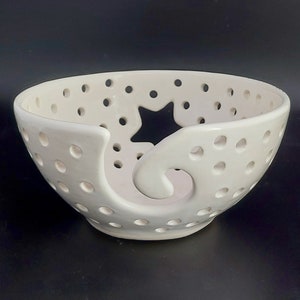 May include: White ceramic yarn bowl with a star-shaped cutout and a swirl design. The bowl has small holes throughout for yarn to pass through.