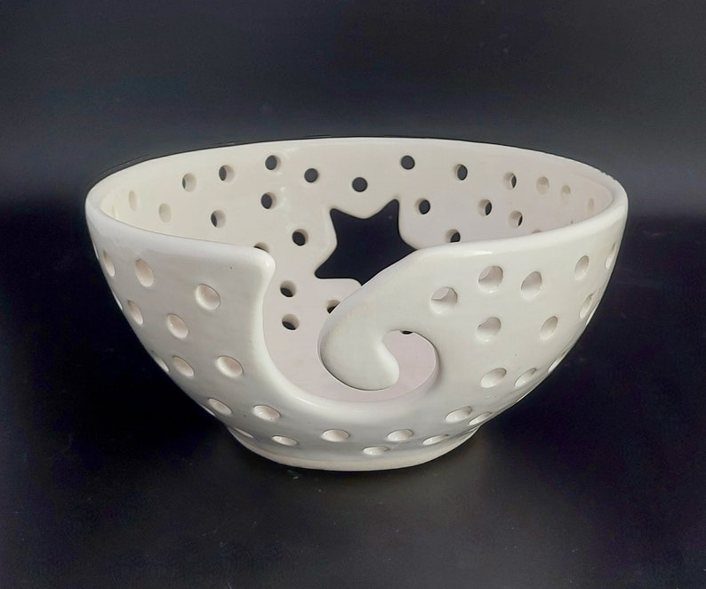 May include: White ceramic yarn bowl with a star-shaped cutout and a swirl design. The bowl has small holes throughout for yarn to pass through.
