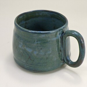 May include: A teal blue ceramic mug with a rounded body and a slightly flared rim. The mug has a handle that is attached to the side of the mug.