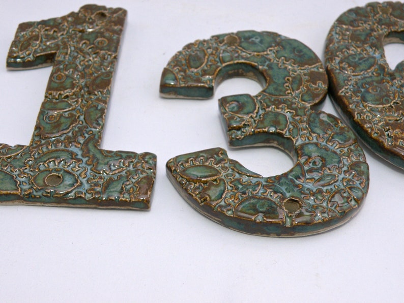 May include: Three ceramic house numbers, 1, 3, and 0, in a green and brown glaze with a decorative pattern. The numbers are individually crafted and have a rustic, handmade look.