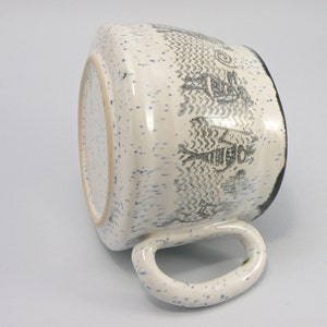 May include: White ceramic mug with a blue and black speckled glaze. The mug has a black rim and a black and white design of stylized figures on the side.