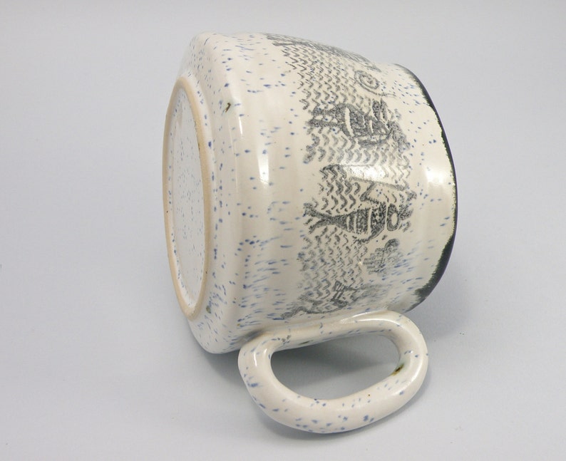 May include: White ceramic mug with a blue and black speckled glaze. The mug has a black rim and a black and white design of stylized figures on the side.