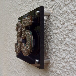 May include: A black acrylic house number sign with the number 35 in raised, textured, green and brown ceramic. The sign is mounted on a white wall with silver screws.