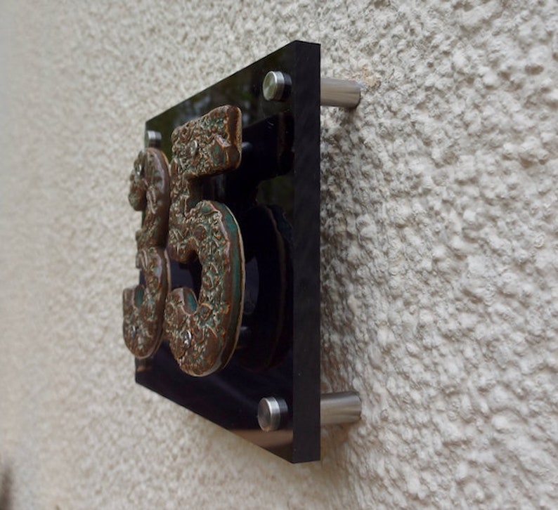 May include: A black acrylic house number sign with the number 35 in raised, textured, green and brown ceramic. The sign is mounted on a white wall with silver screws.