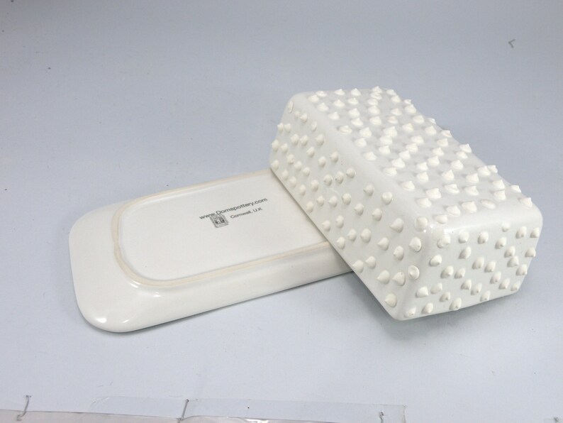 Puede incluir: Una mantequera de cer&aacute;mica blanca con base rectangular y tapa cubierta de peque&ntilde;os detalles c&oacute;nicos en relieve. La mantequera est&aacute; sobre una bandeja rectangular a juego. El texto "www.domspottery.com" es visible en la bandeja.