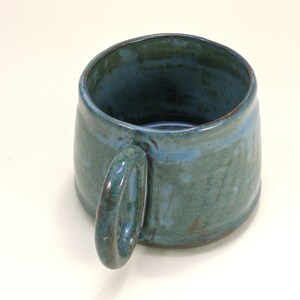 May include: A handmade ceramic mug with a teal glaze and a brown handle. The mug has a rustic, handmade look.