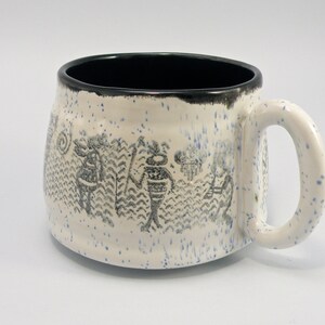 May include: White ceramic mug with a black rim and a design of stylized figures in black on the side. The mug has a speckled glaze.
