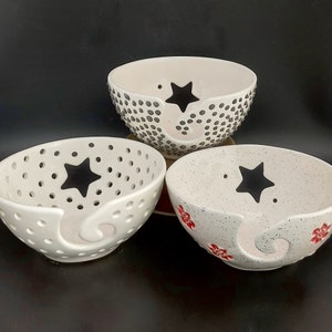 May include: Three white ceramic yarn bowls with star designs. The bowls have a hole on the side for yarn to feed through. One bowl has black polka dots, one has black stars, and one has red flowers.