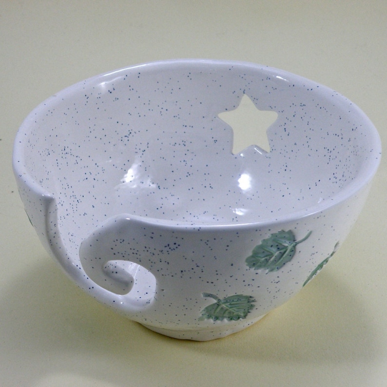 Puede incluir: Cuenco de lana de cer&aacute;mica blanca con un agujero en forma de estrella y dise&ntilde;os de hojas verdes. El cuenco tiene una gu&iacute;a de lana incorporada.