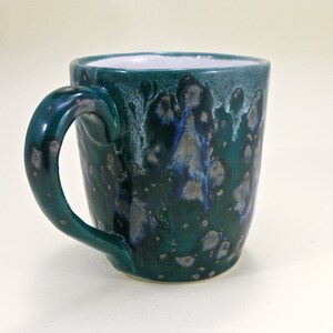 May include: A teal green ceramic mug with a white interior and a speckled glaze. The mug has a unique, asymmetrical handle.