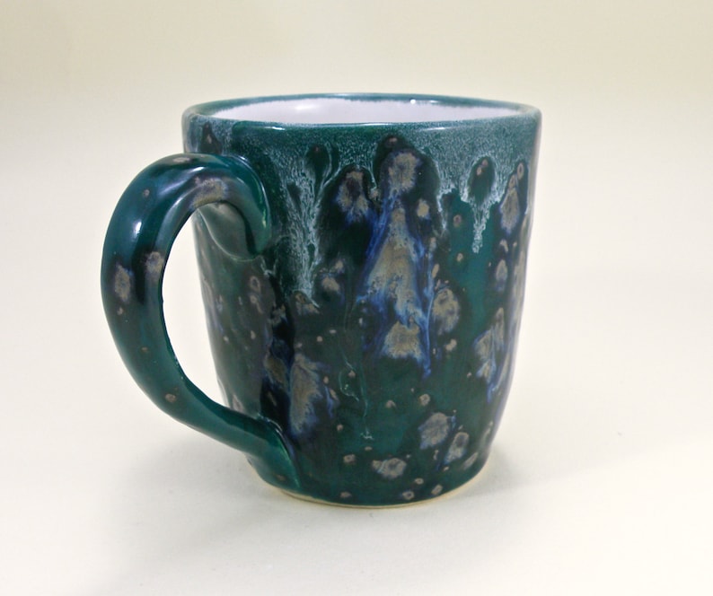 May include: A teal green ceramic mug with a white interior and a speckled glaze. The mug has a unique, asymmetrical handle.