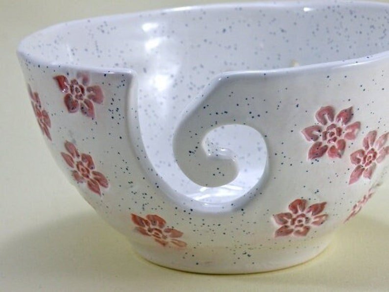 May include: White ceramic yarn bowl with a pink floral design and a large, curved opening for yarn.