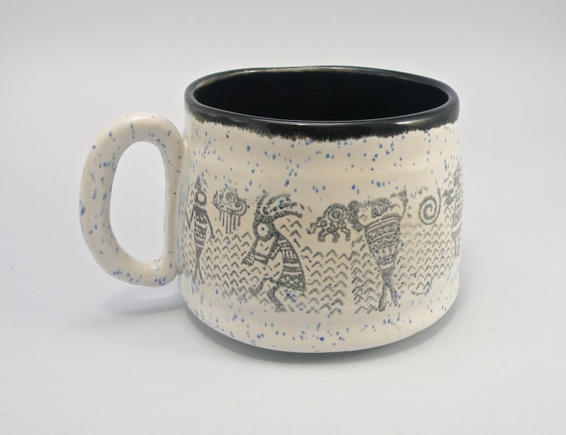May include: White ceramic mug with a black rim and a blue and black design of stylized figures. The mug has a speckled blue glaze.