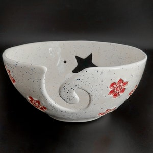 May include: White ceramic yarn bowl with a star and red flower designs. The bowl has a hole in the side for yarn to feed through.