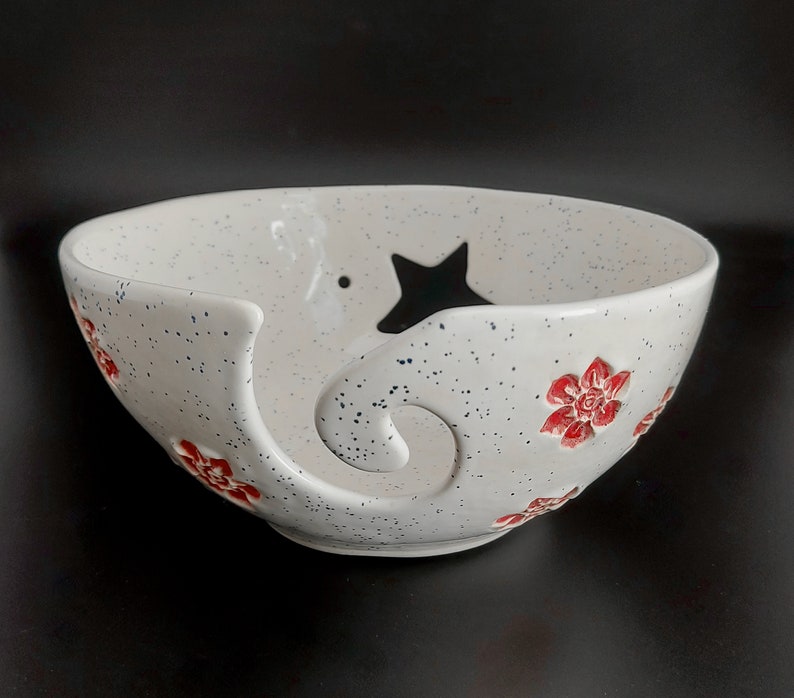 May include: White ceramic yarn bowl with a star and red flower designs. The bowl has a hole in the side for yarn to feed through.