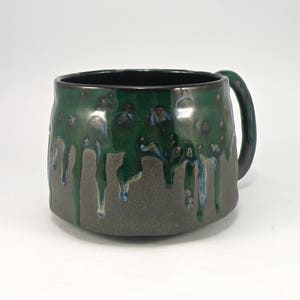 May include: A dark green and black ceramic mug with a drip glaze design. The mug has a rounded body and a handle.