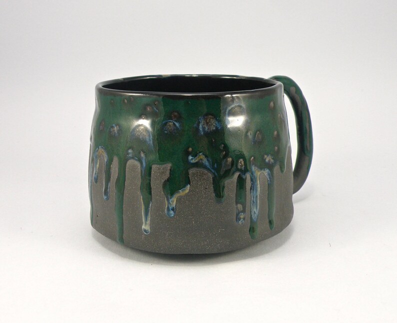 May include: A dark green and black ceramic mug with a drip glaze design. The mug has a rounded body and a handle.