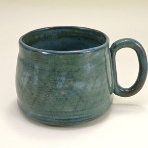 May include: A teal blue ceramic mug with a rounded body and a slightly flared rim. The mug has a speckled glaze and a brown handle.