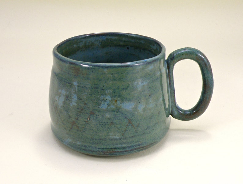 May include: A teal blue ceramic mug with a rounded body and a slightly flared rim. The mug has a speckled glaze and a brown handle.
