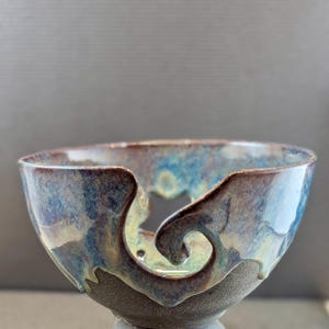 May include: A ceramic yarn bowl with a blue, green, and brown glaze. The bowl has a unique design with a cutout for yarn to pass through.