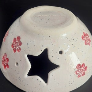 May include: White ceramic bowl with a star-shaped cutout and red floral designs. The bowl has a speckled glaze and a white rim.