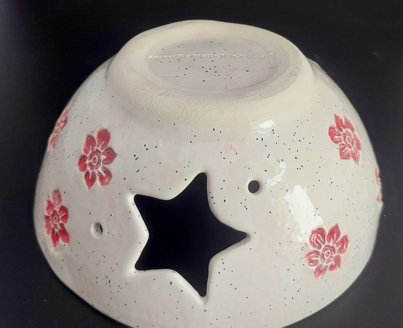 May include: White ceramic bowl with a star-shaped cutout and red floral designs. The bowl has a speckled glaze and a white rim.