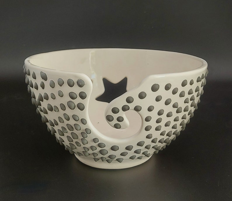 May include: White ceramic yarn bowl with a star and a swirl design. The bowl is covered in small gray dots.