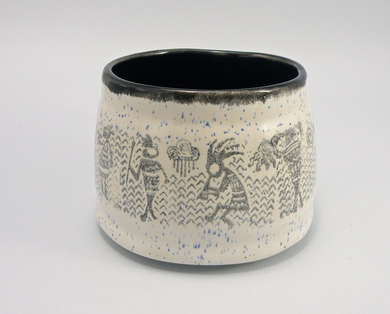 May include: White ceramic bowl with a black rim and a speckled glaze. The bowl features a black and white design of stylized figures and a rain cloud.