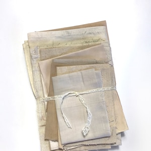 May include: A stack of assorted fabric swatches in neutral tones, including beige, tan, and cream. The fabrics are tied together with a delicate white lace ribbon. The textures vary from linen to rougher weaves.