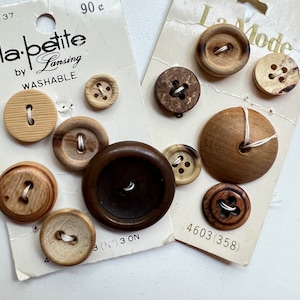 May include: Two vintage cardboard display cards featuring assorted round wooden buttons in various shades of brown. The left card is labeled la petite by Lansing and the right card is labeled La Mode. The buttons vary in size, texture, and grain.