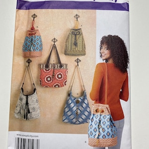 May include: Simplicity sewing pattern 1598 os for five different bags. The bags are shown hanging on hooks and being worn by a model. The bags are made of different fabrics and have different designs. The pattern includes instructions for making a drawstring bag, a tote bag, a hobo bag, and two other styles of bags.