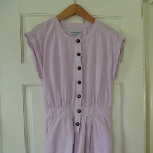 May include: A lavender button-up jumpsuit with short sleeves and a round neckline. The jumpsuit has a brand tag that reads "Urban Outfitters".