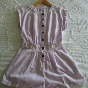 May include: A light purple linen jumpsuit with a button-up front and short sleeves. The jumpsuit has a relaxed fit and a slightly gathered waist.