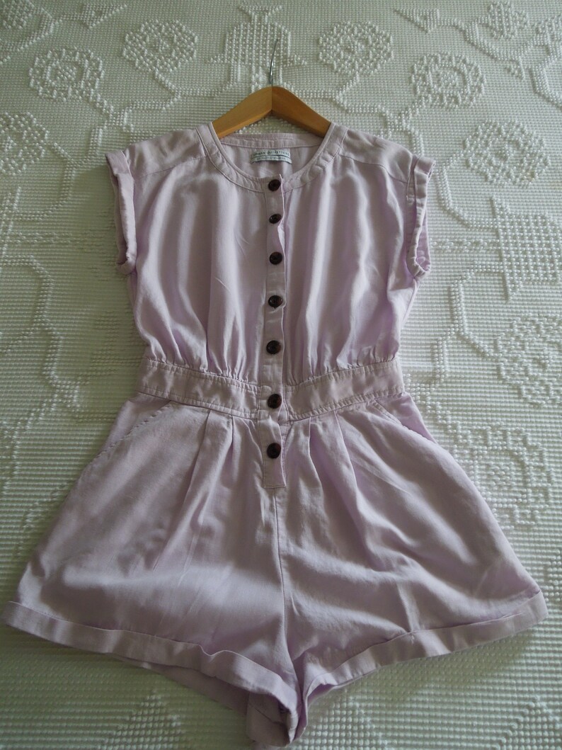 May include: A light purple linen jumpsuit with a button-up front and short sleeves. The jumpsuit has a relaxed fit and a slightly gathered waist.